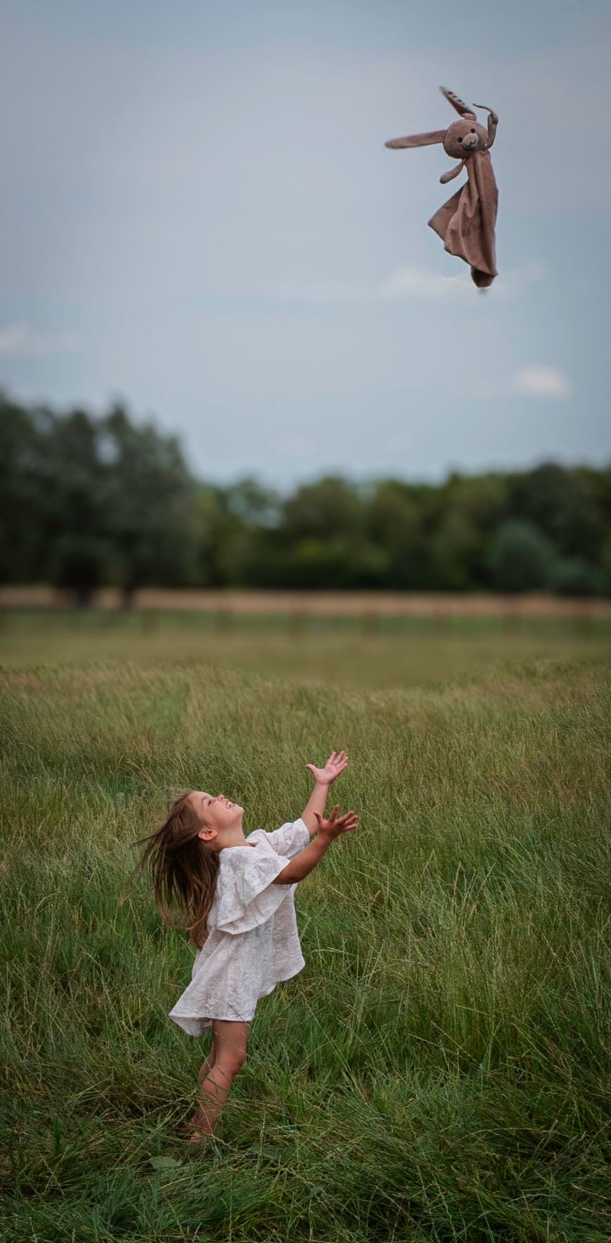Young girl in white dress tossing a stuffed bunny toy high into the air in an open field.
