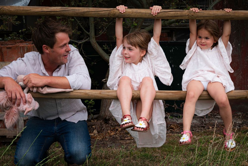 Father watching two young daughters hanging and swinging on a rustic wooden fence outdoors.
