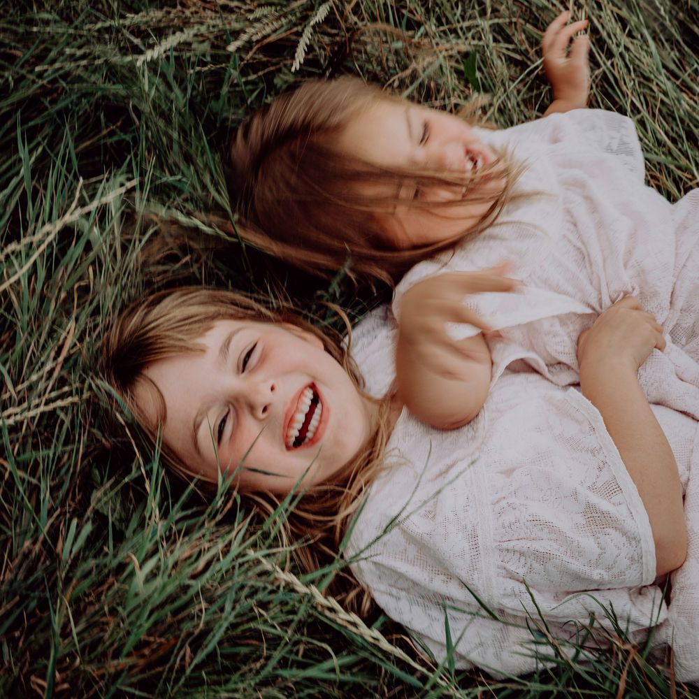 Two young sisters laughing and lying together in tall green grass during an outdoor photo session.