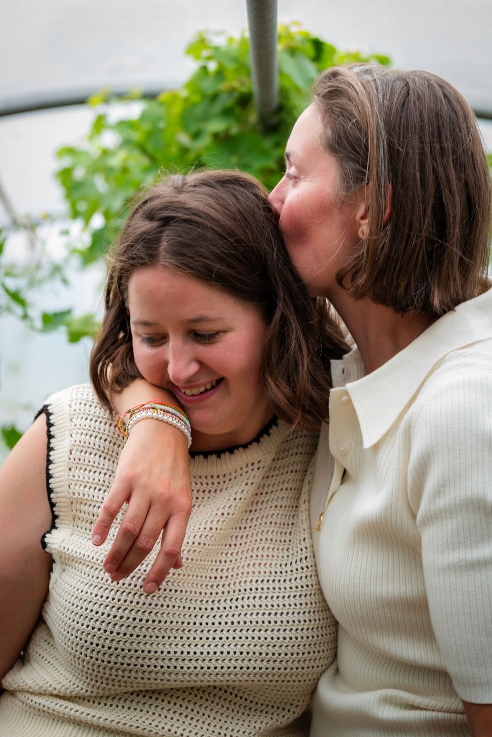 Two women sharing a tender moment, one kissing the other on the cheek while embracing in a greenhouse setting.