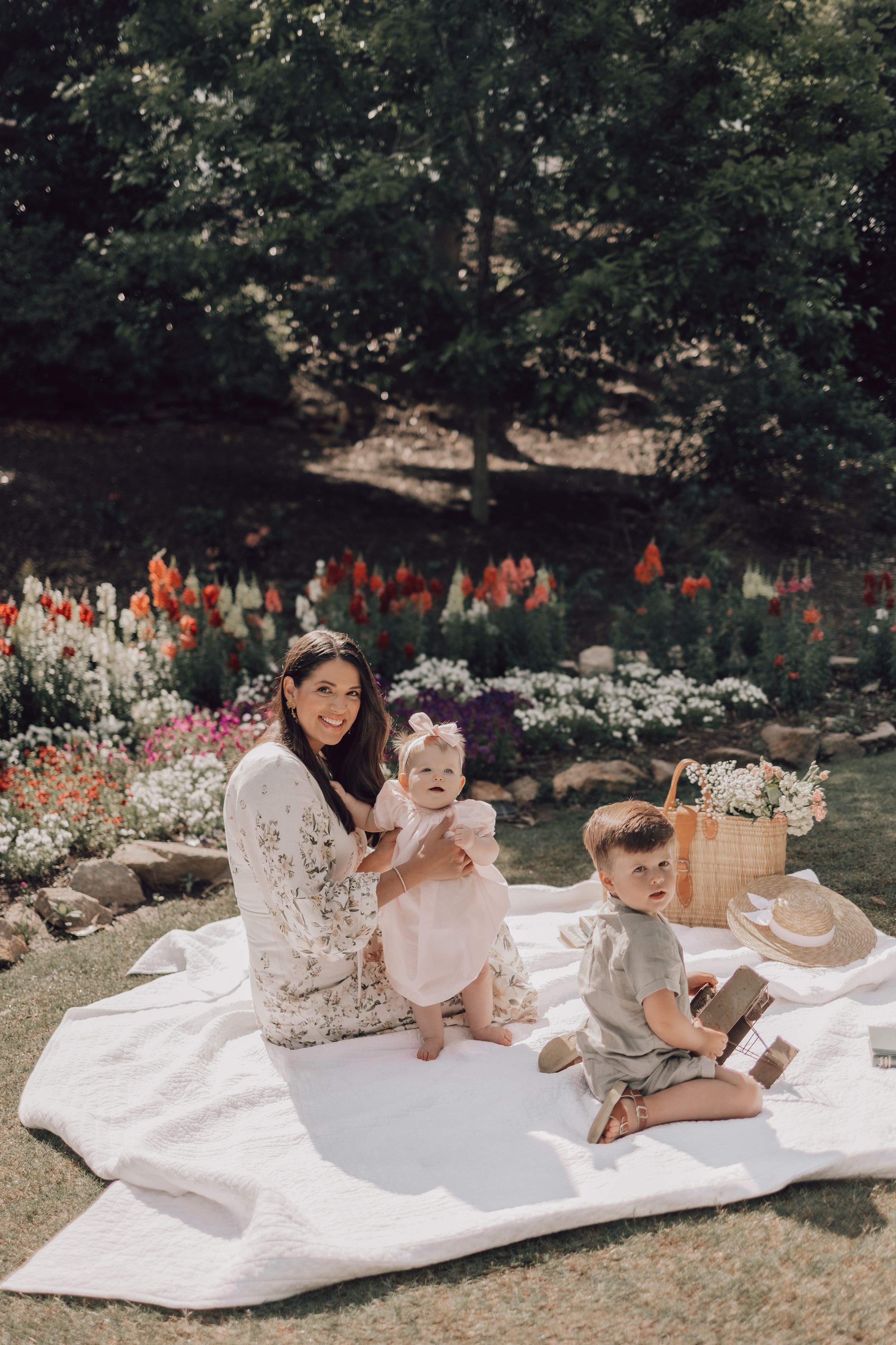 Family relaxing together on white blanket in garden with red and white flowers in background.