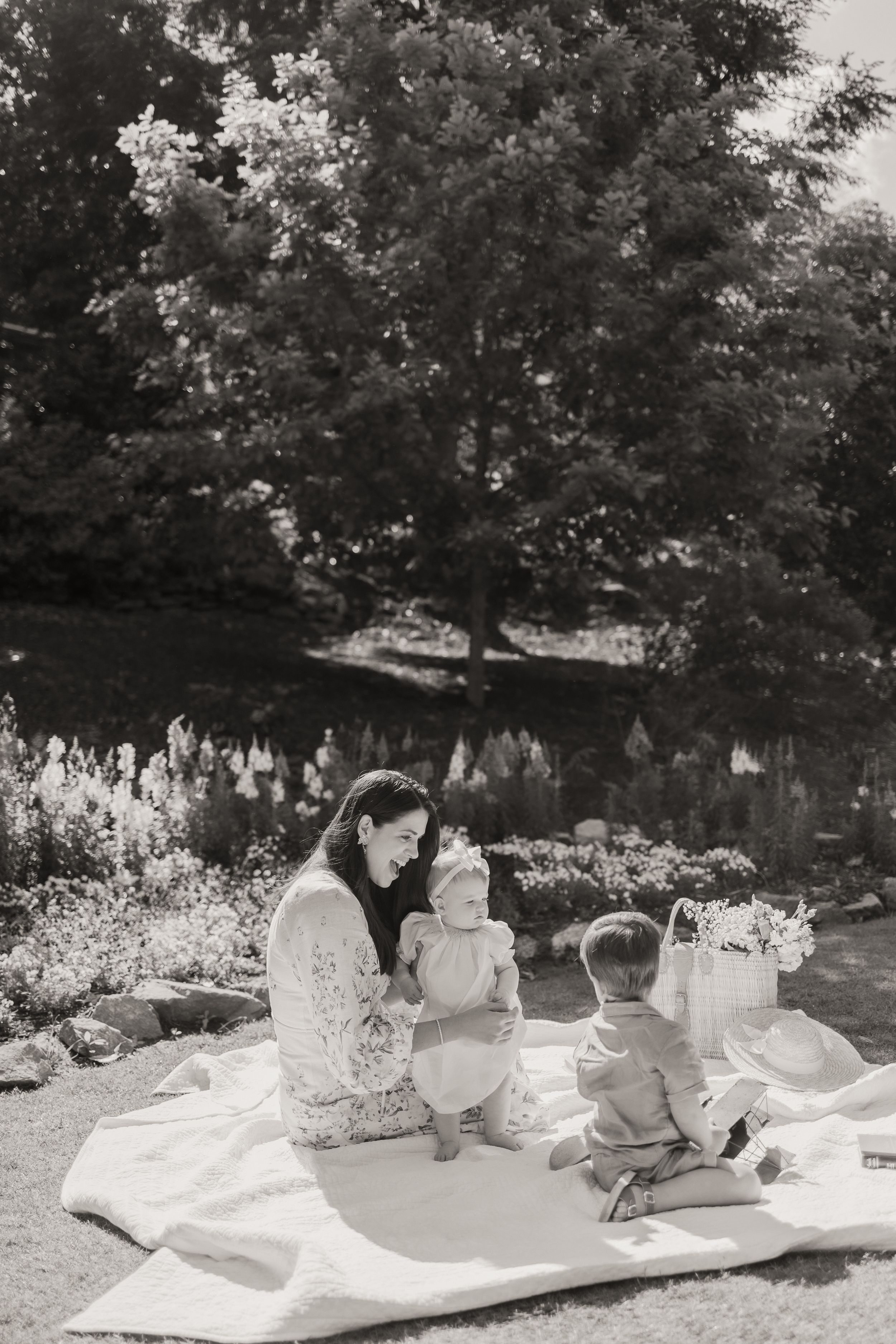 Three children lying on a white blanket outdoors reading books together in a garden setting.
