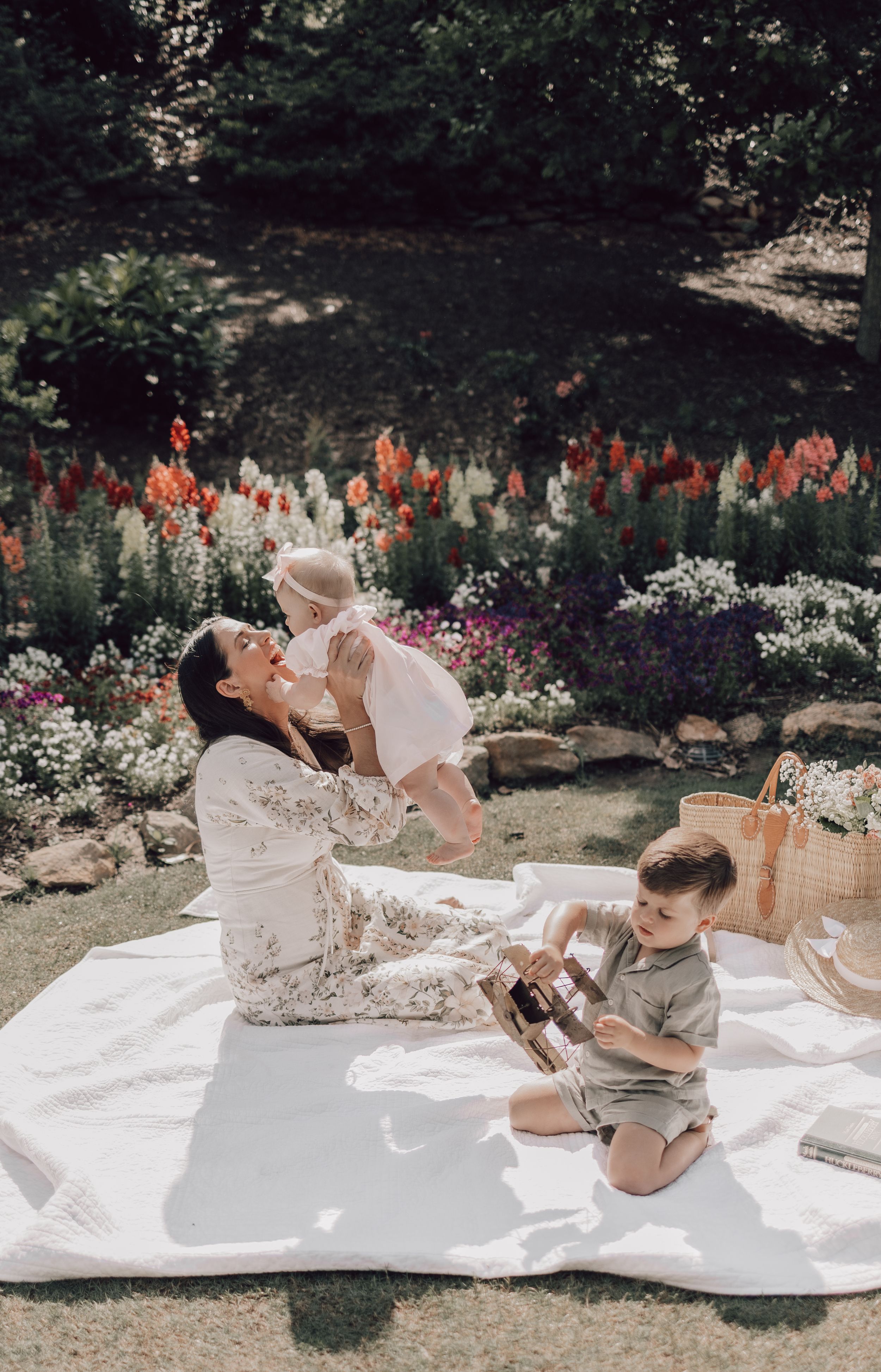 A family enjoys a playful moment together on a white blanket in a garden with colorful flowers in the background.