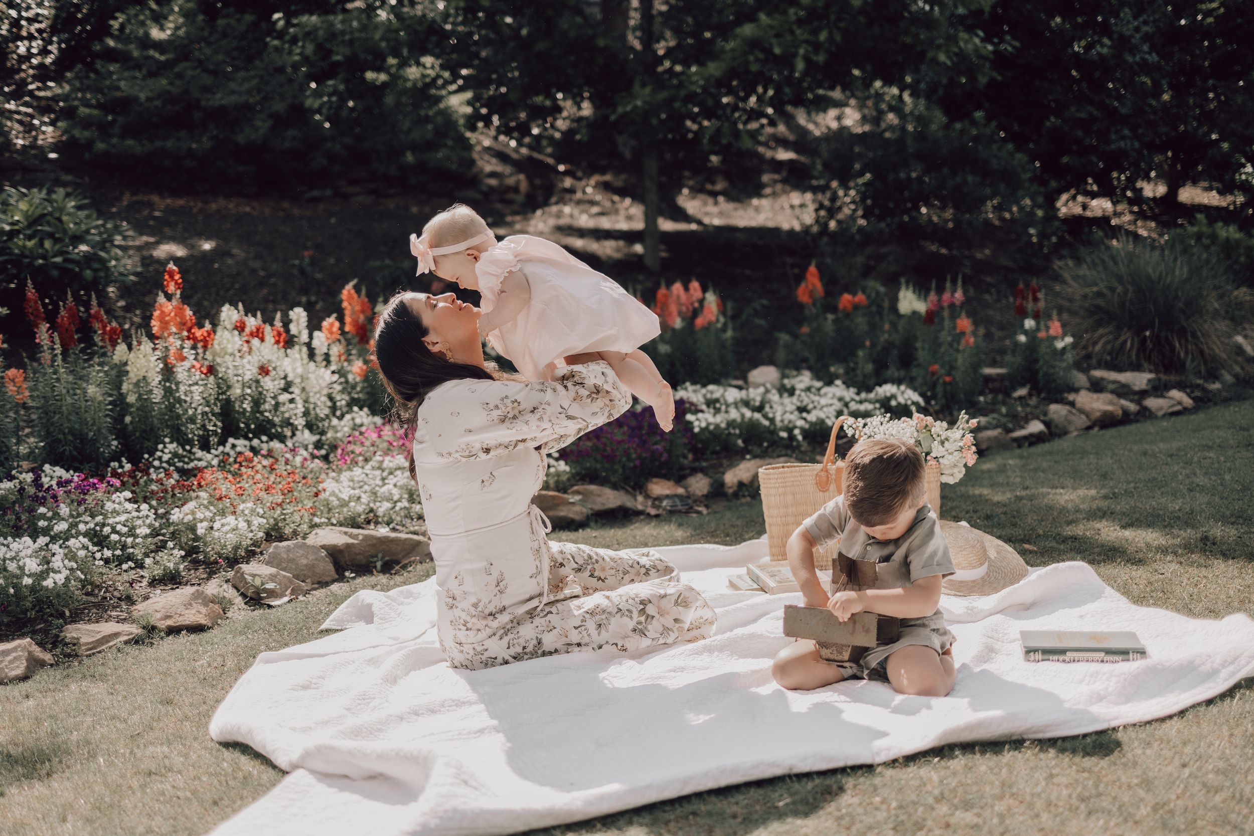 Two people in wedding attire sit together outdoors in a garden setting with flowers and greenery around them.