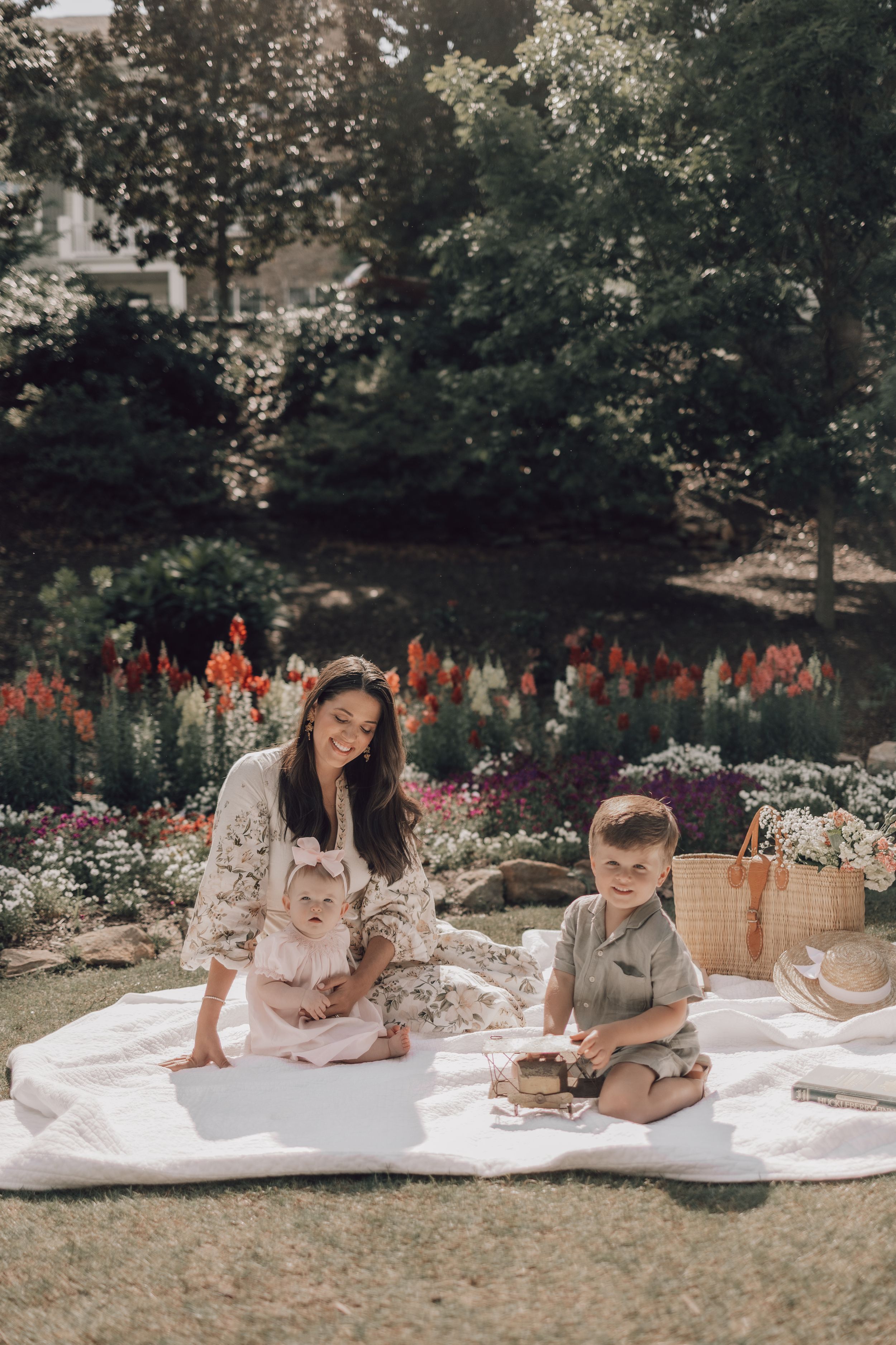 Two children sit on a white blanket in a garden setting doing an activity together while surrounded by flowers.