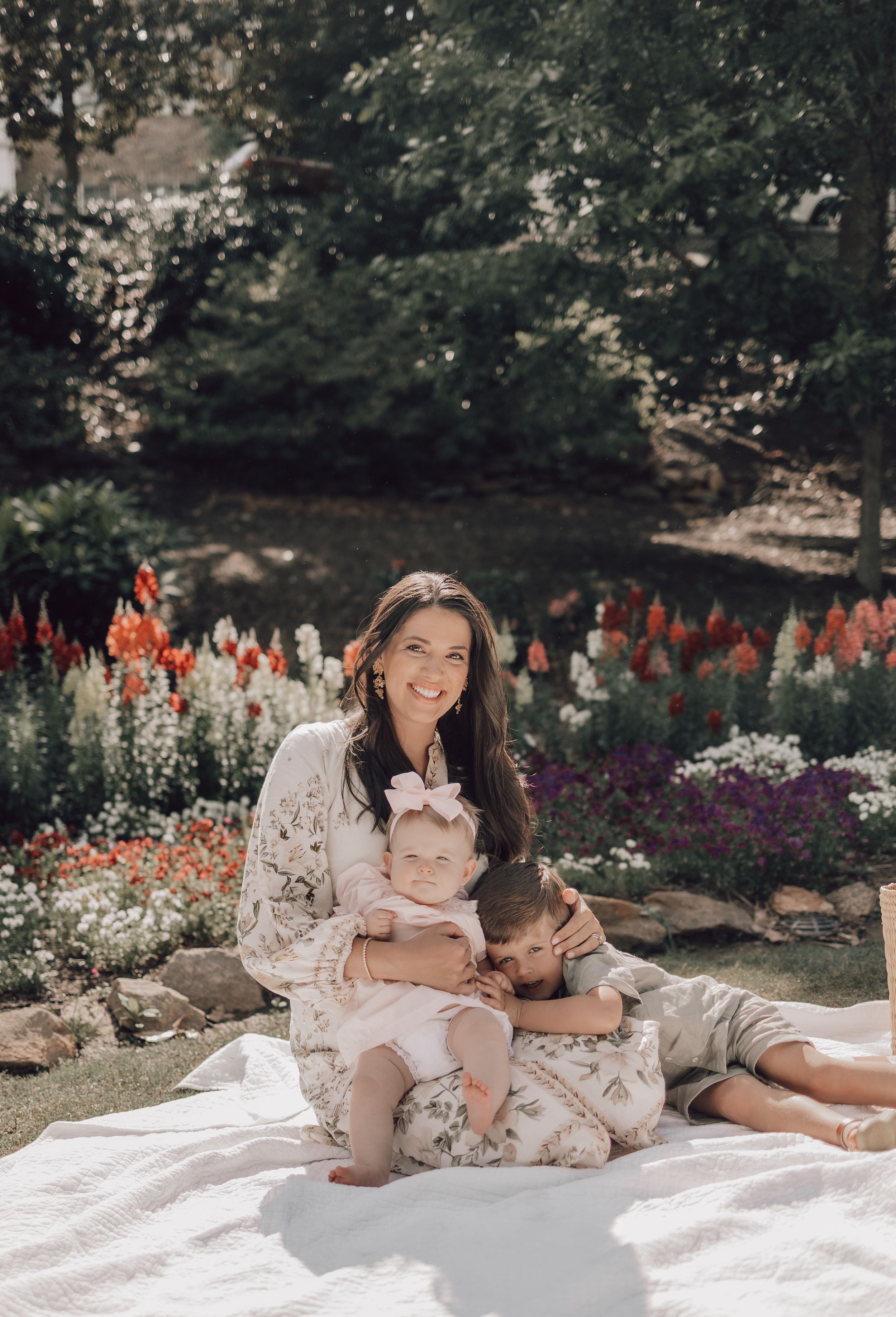Family enjoying a summer picnic on white blanket in garden with colorful flowers in background.