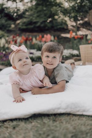 Two children lying on a white blanket in a garden with flowers in the background.