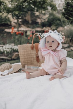 A baby in a pink bonnet and dress sits on a white blanket with a picnic basket in a garden setting.