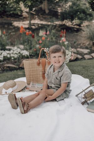 A child relaxes on a white blanket in a garden setting with a picnic basket and flowers in the background.