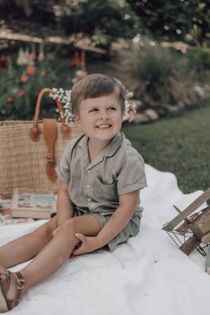 A child in a grey shirt lies on a white blanket outdoors with a woven basket nearby.