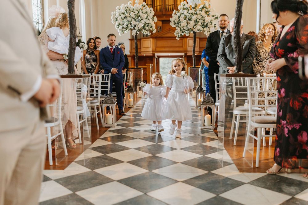 A flower girl in a white dress walks down a black and white checkered aisle at an elegant wedding ceremony.