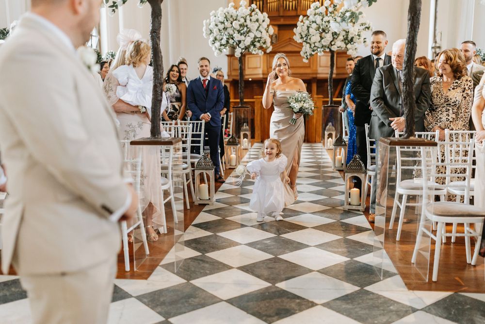 A wedding ceremony takes place in an elegant venue with a black and white checkered floor and white floral arrangements.