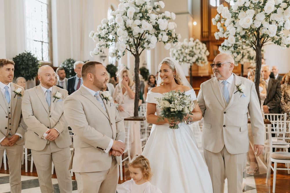 Wedding party in light gray suits and white dresses pose together in elegant reception venue with floral decorations.