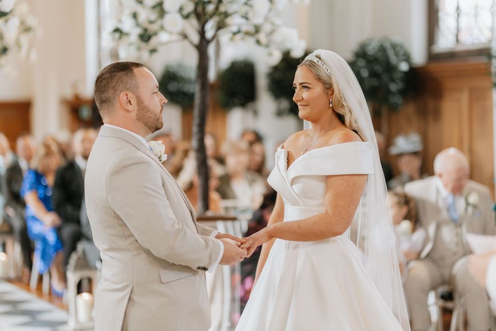A romantic wedding ceremony moment in a bright indoor venue with white decor and guests seated in the background.