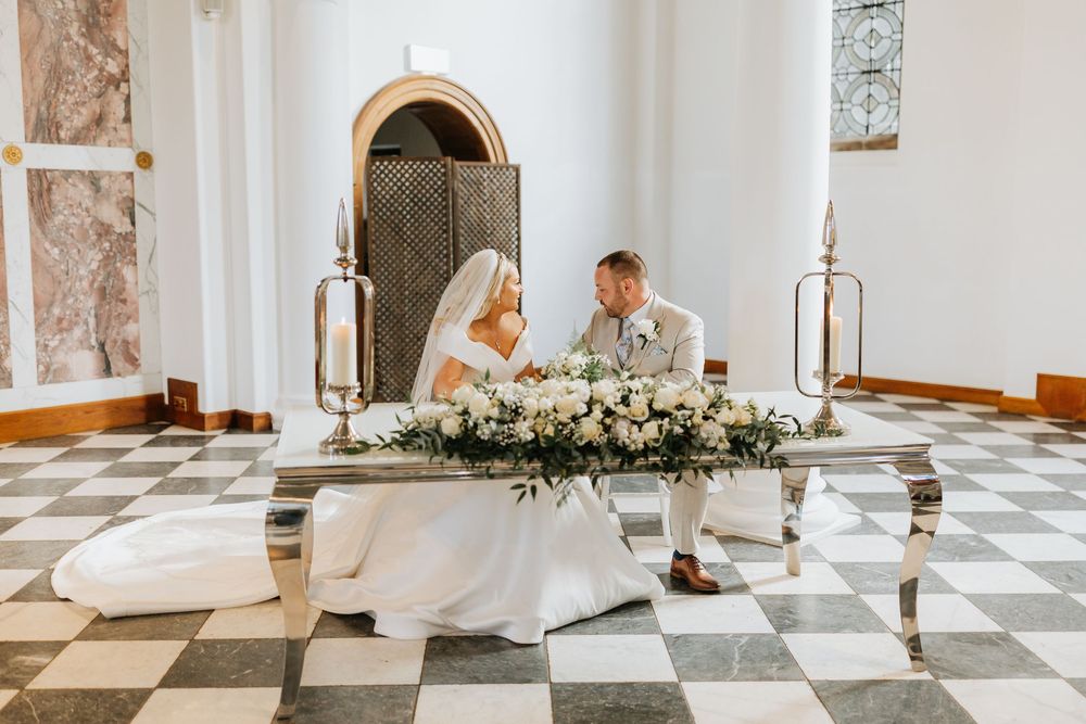 An elegant wedding reception table with white floral centerpieces and checkerboard flooring in a light-filled venue.