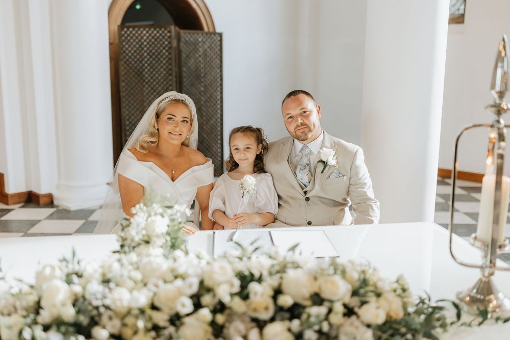 Wedding party sits at white-decorated table with floral arrangements in bright indoor venue.