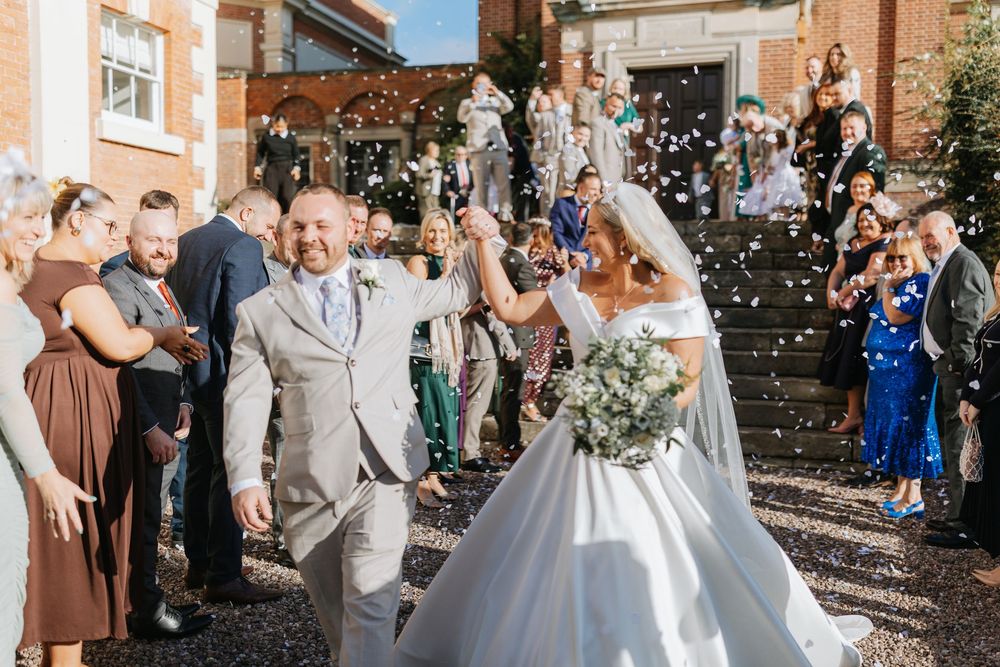 Wedding party celebrates with confetti toss outside brick building during autumn ceremony.