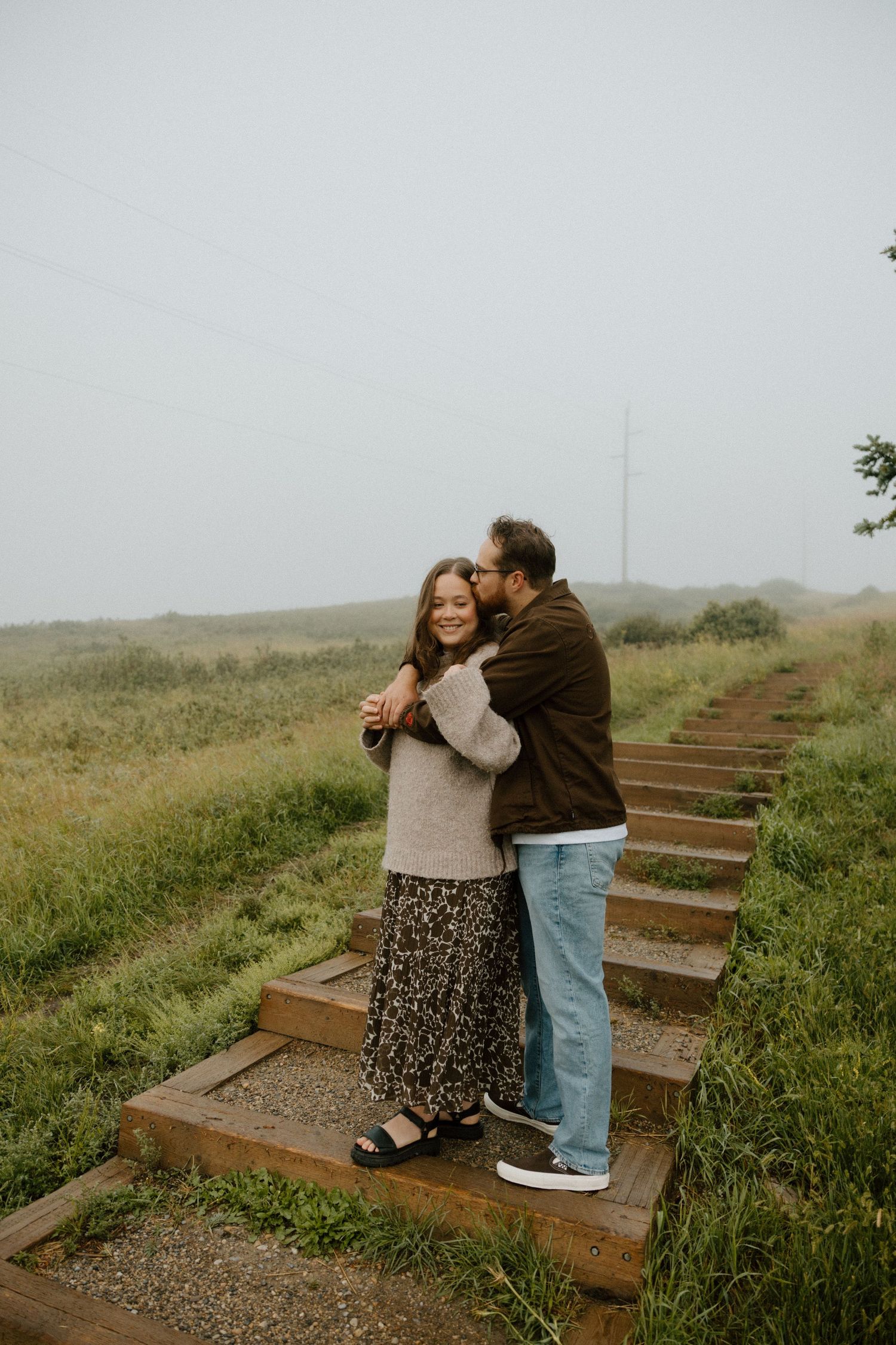 A couple embraces on wooden steps along a misty coastal trail surrounded by green vegetation.