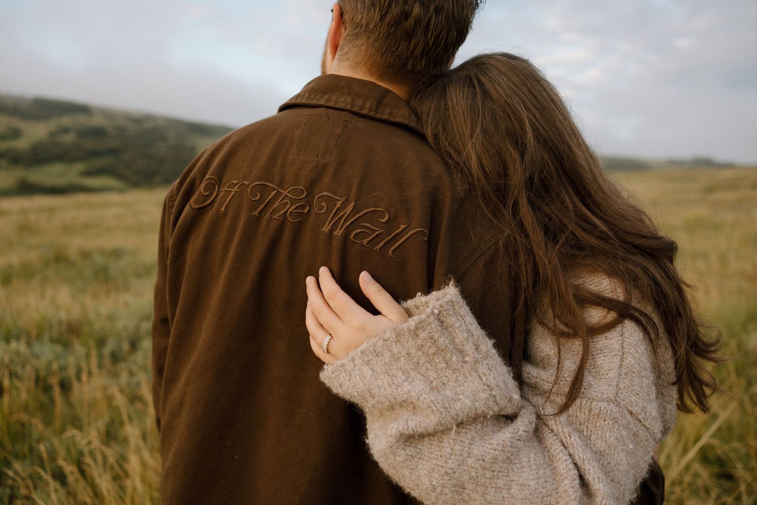 A couple shares an intimate moment in a brown jacket and sweater against a rural landscape.
