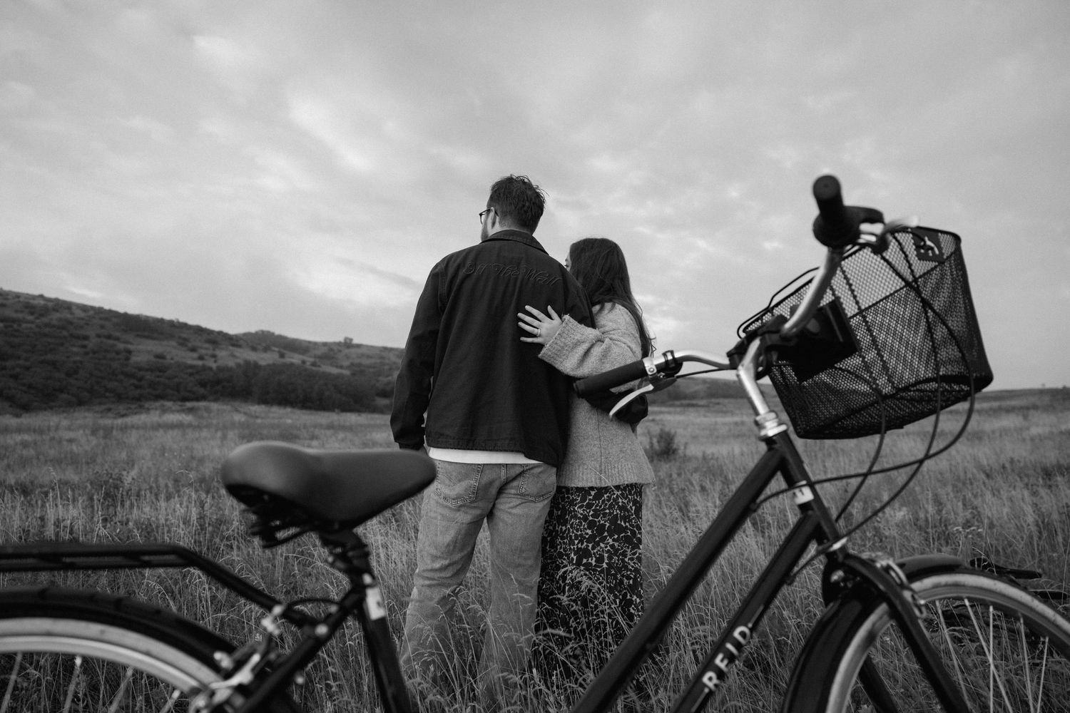 A couple embraces next to a bicycle with a basket on a scenic trail at dusk.