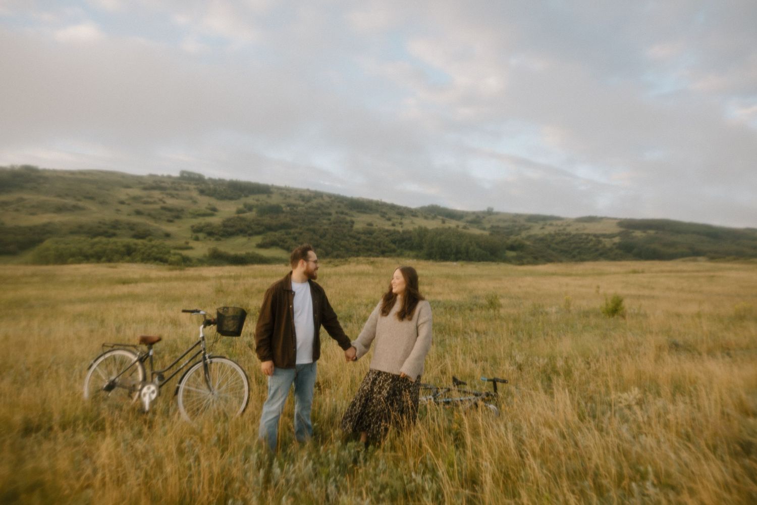 Two vintage bicycles rest in tall grass with rolling hills in the background.