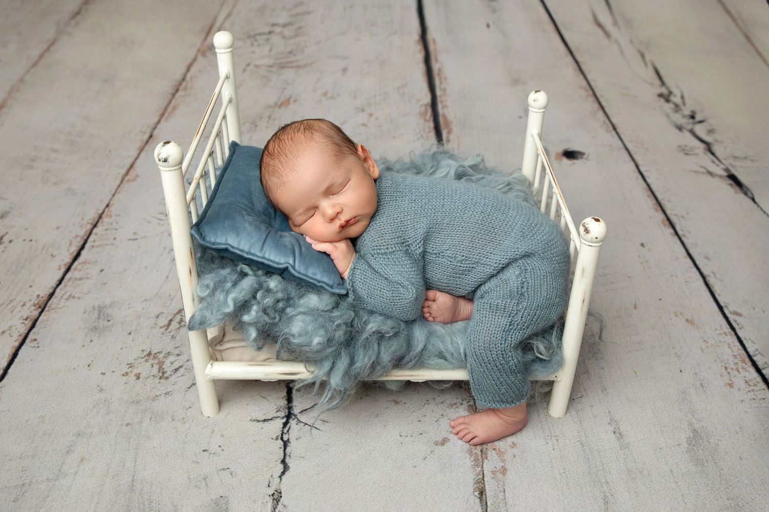 Newborn baby sleeping peacefully in a tiny white bed with blue knit outfit and fluffy blanket in a professional photo.