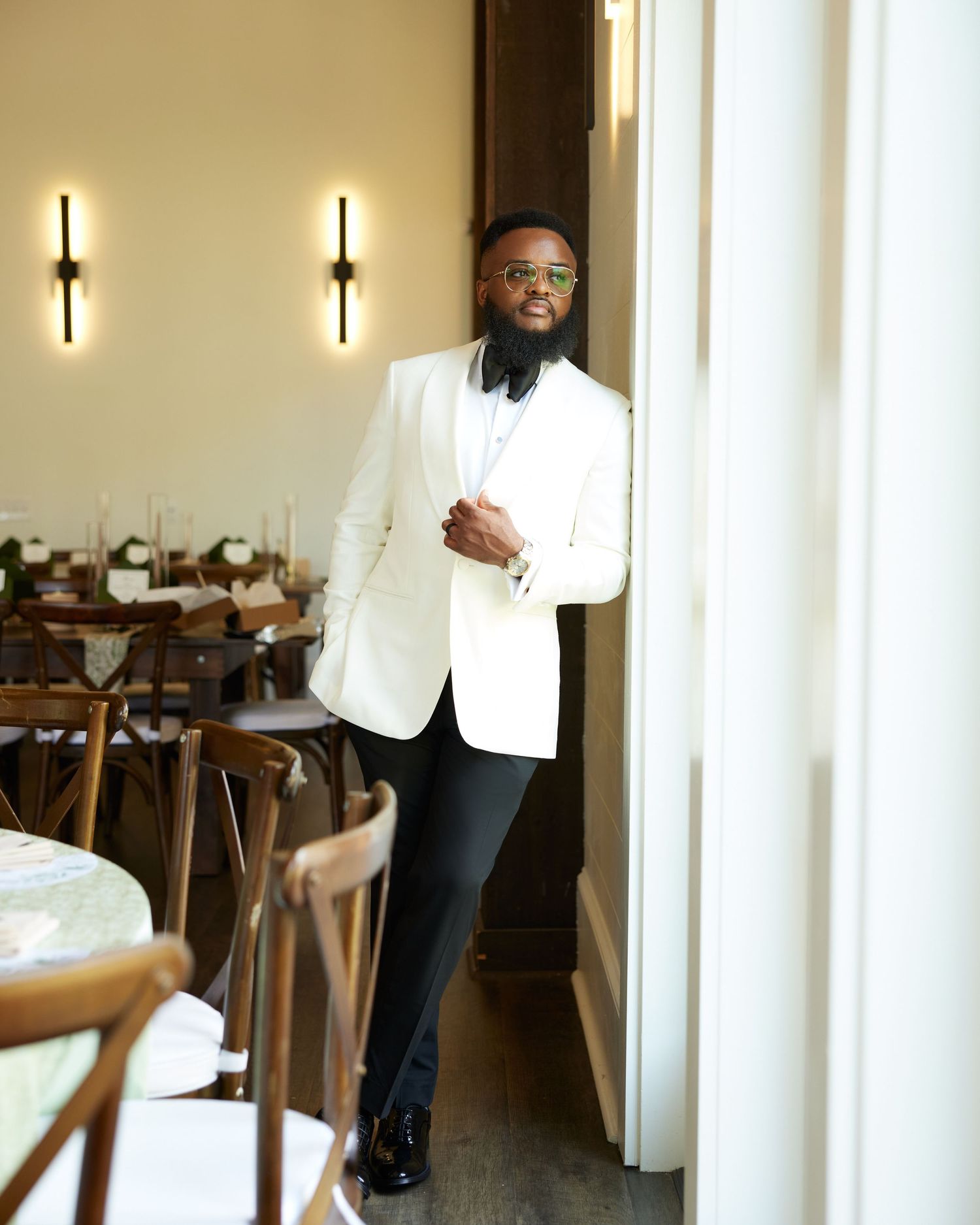 Stylish man in white blazer and black pants posing elegantly in a modern restaurant setting.