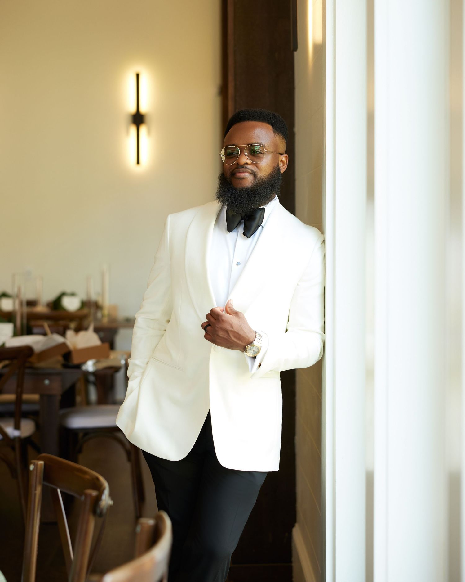 A stylish man in a white tuxedo jacket and black bow tie poses confidently in an elegant restaurant setting.