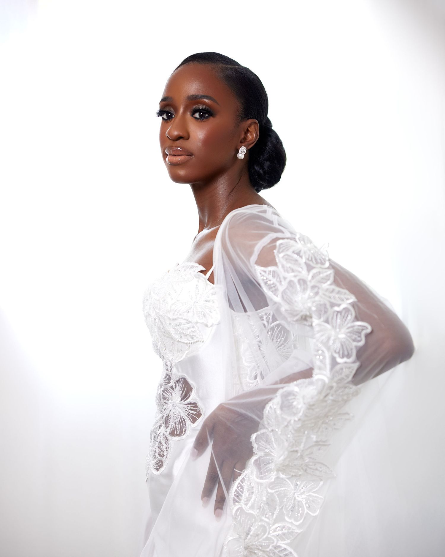 An elegant bride wearing a white lace wedding gown with sheer floral cape sleeves poses gracefully.