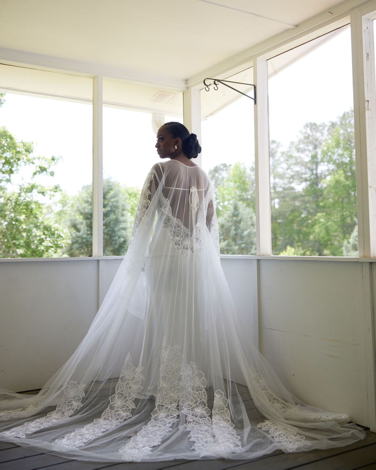 Bride in elegant white lace wedding gown with sheer cape train stands before sunlit windows overlooking green trees.