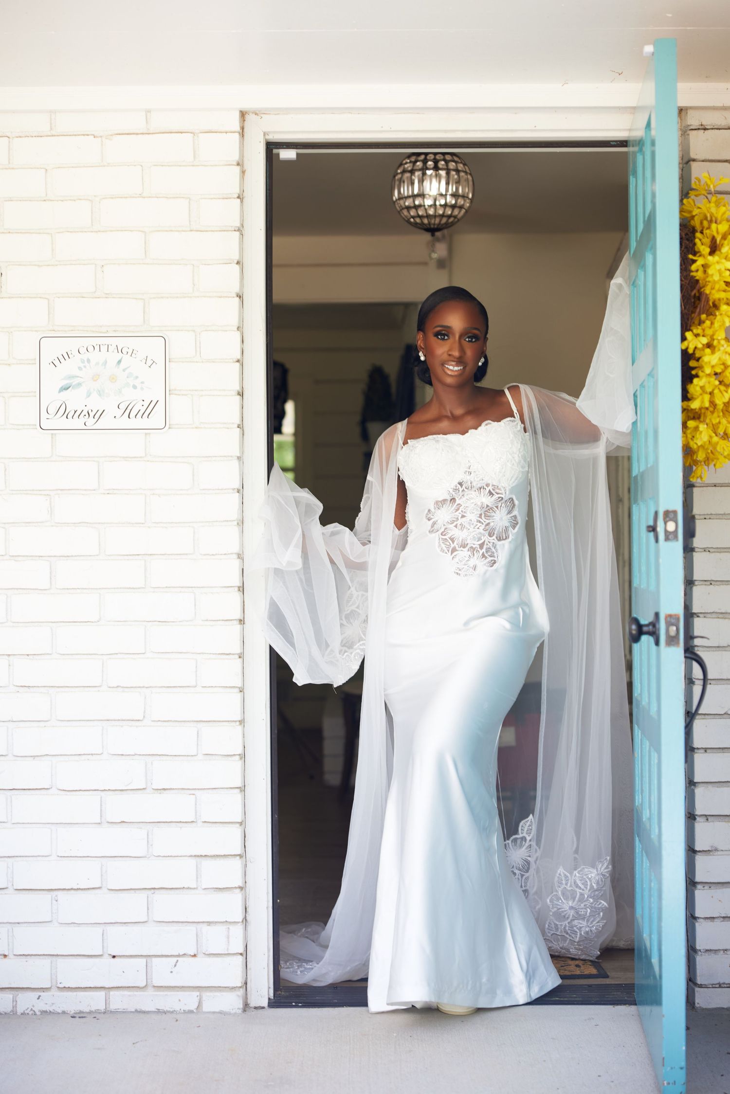 Smiling bride in white mermaid gown with sheer cape stands in doorway of The Cottage at Daisy Hill.