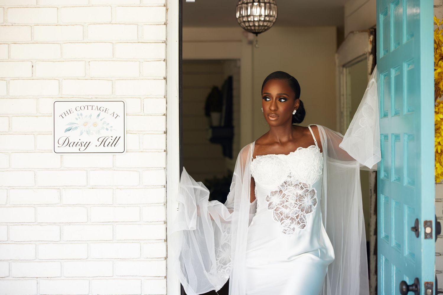 Elegant bride in white wedding gown and sheer wrap stands in doorway at Daisy Hill cottage.