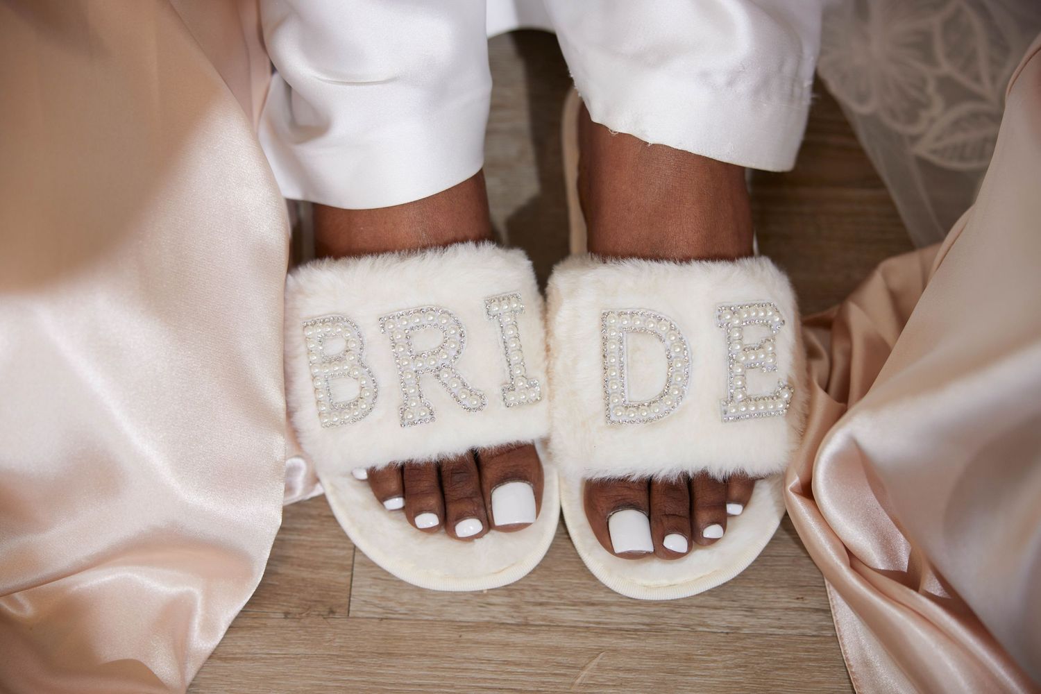 Close-up of a bride wearing fluffy white BRIDE slippers with rhinestone letters, showing white pedicured toes.