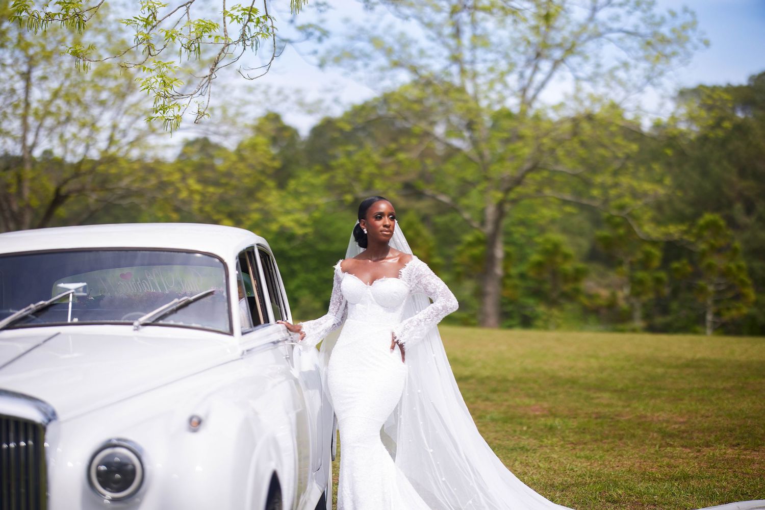 Bride in white mermaid gown with long veil poses beside a classic white vintage car on a green lawn.