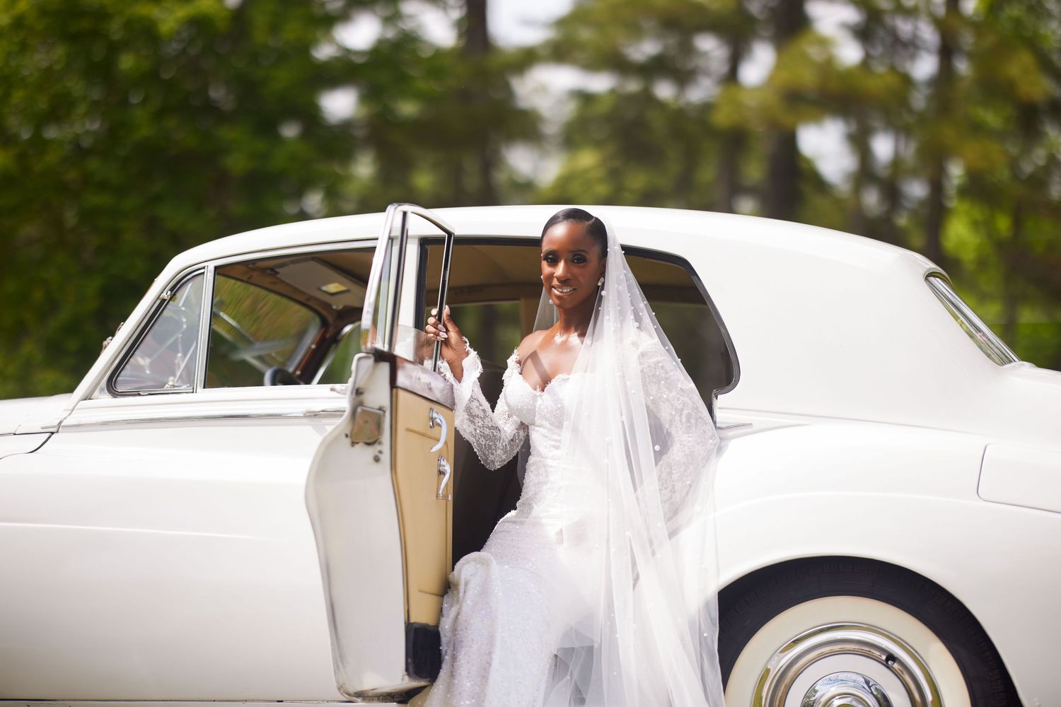 Elegant bride in white wedding dress and veil posing by a classic white car on her wedding day.