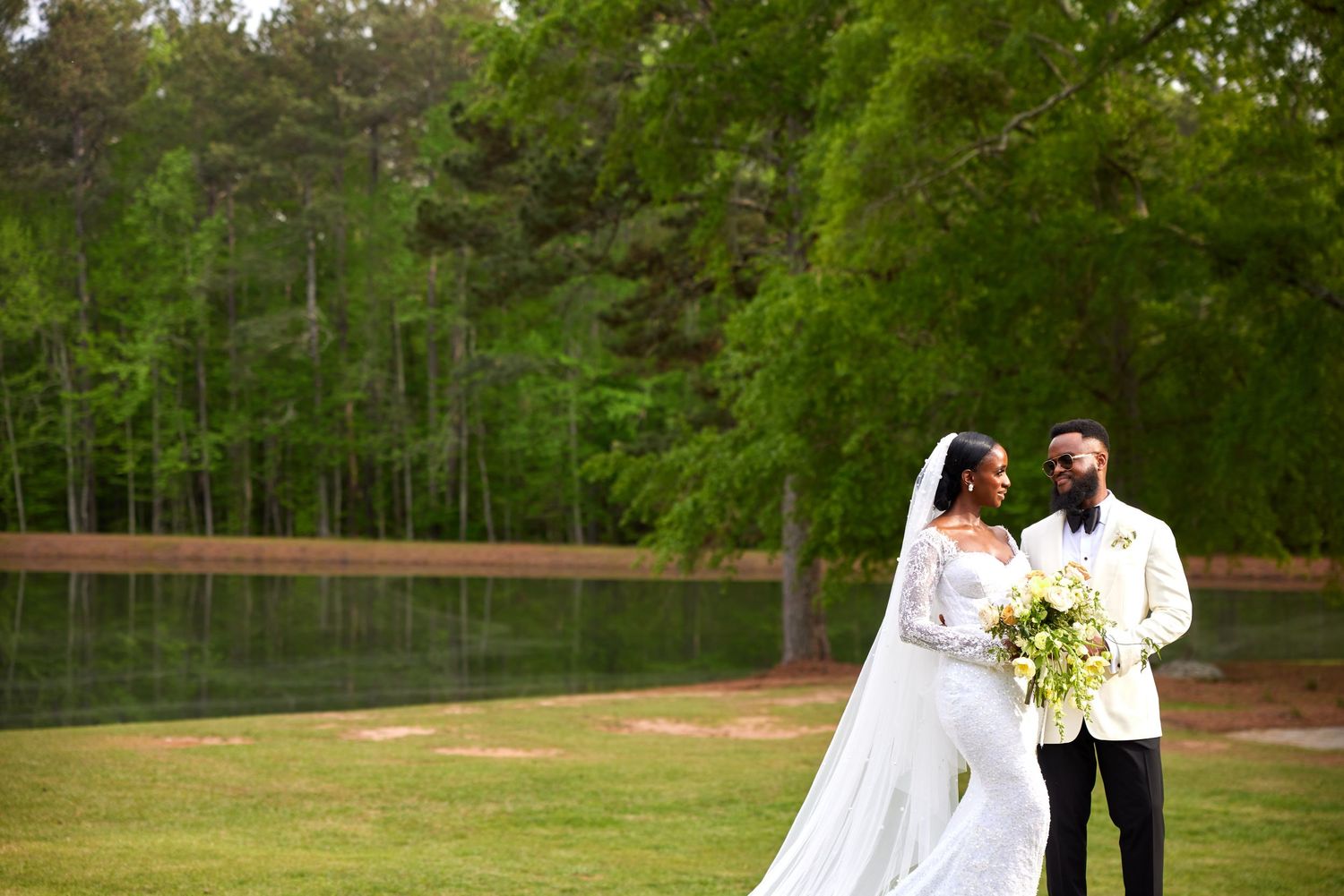 Elegant bride and groom pose on lush green lawn near pond, bride in white gown with veil, groom in white tuxedo.