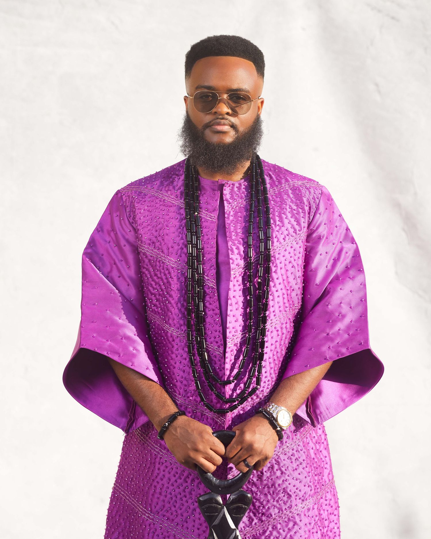 Man in purple African agbada outfit with black beaded necklace and sunglasses posing confidently.