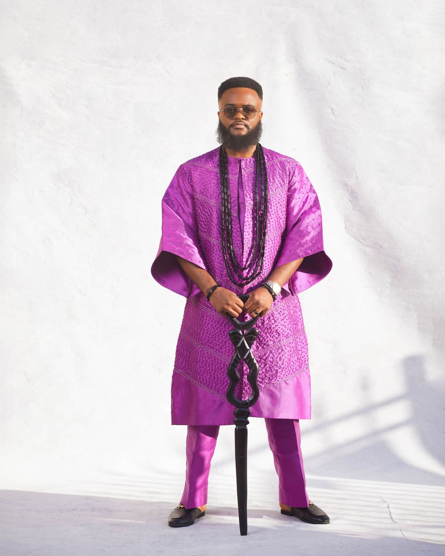 Man in vibrant purple African agbada outfit holding a decorative staff, posing against a white wall.