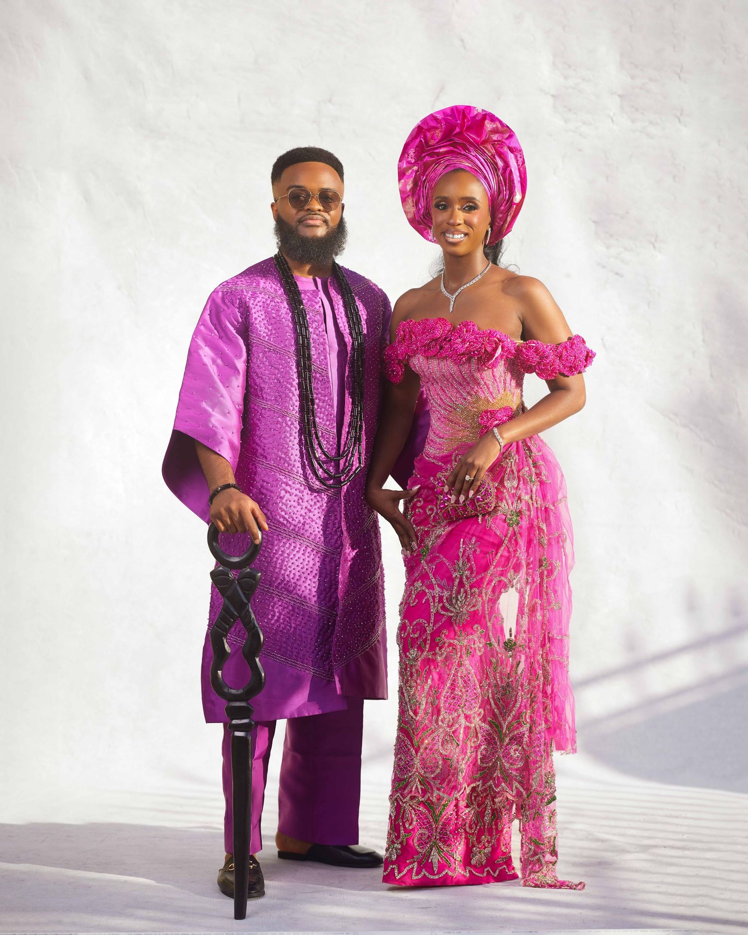 Couple in matching vibrant pink Nigerian traditional attire, man in agbada and woman in lace gown with gele headwrap.