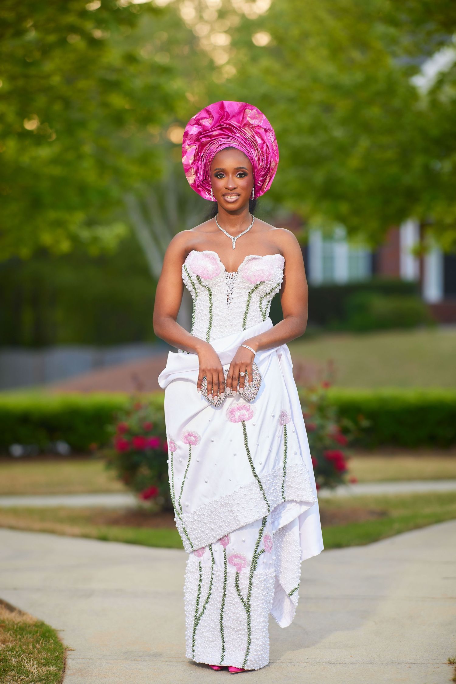 Woman in white floral gown and pink Nigerian gele headwrap poses outdoors in elegant traditional African bridal attire.