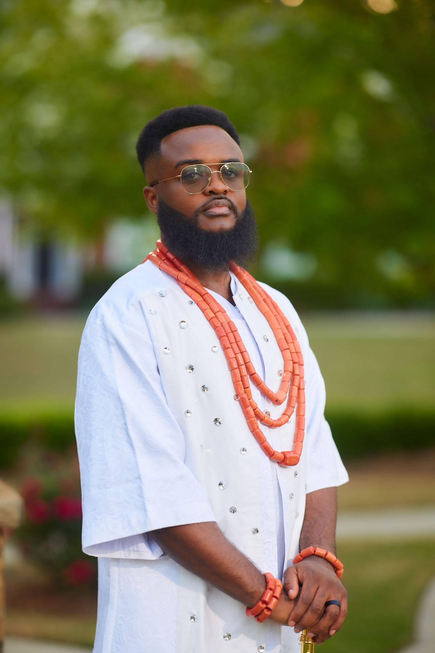 Man in white traditional Nigerian attire with coral beaded necklaces and sunglasses posing outdoors.