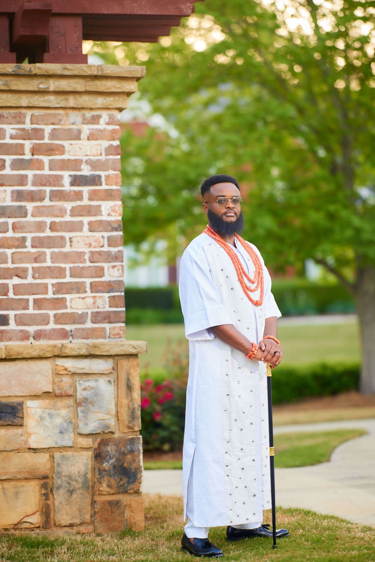 Man in white traditional African attire with orange coral beads stands beside a brick pillar outdoors.
