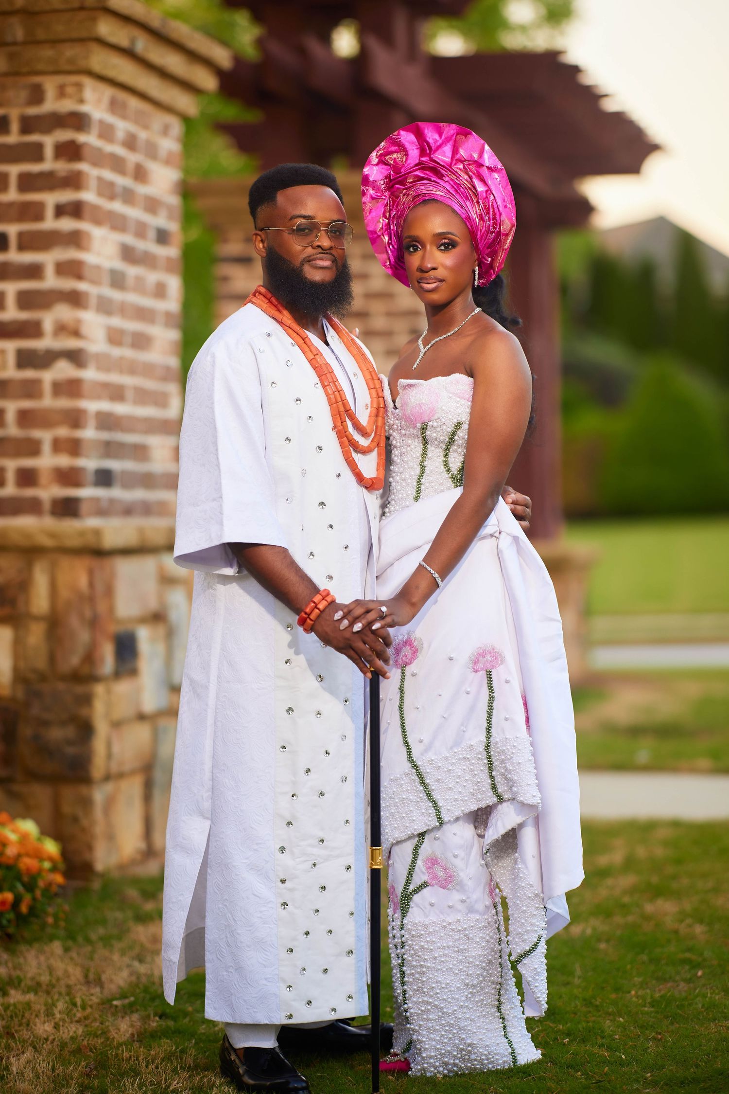 Couple in traditional Nigerian wedding attire, man in white agbada with coral beads, woman in white gown with pink gele headwrap.