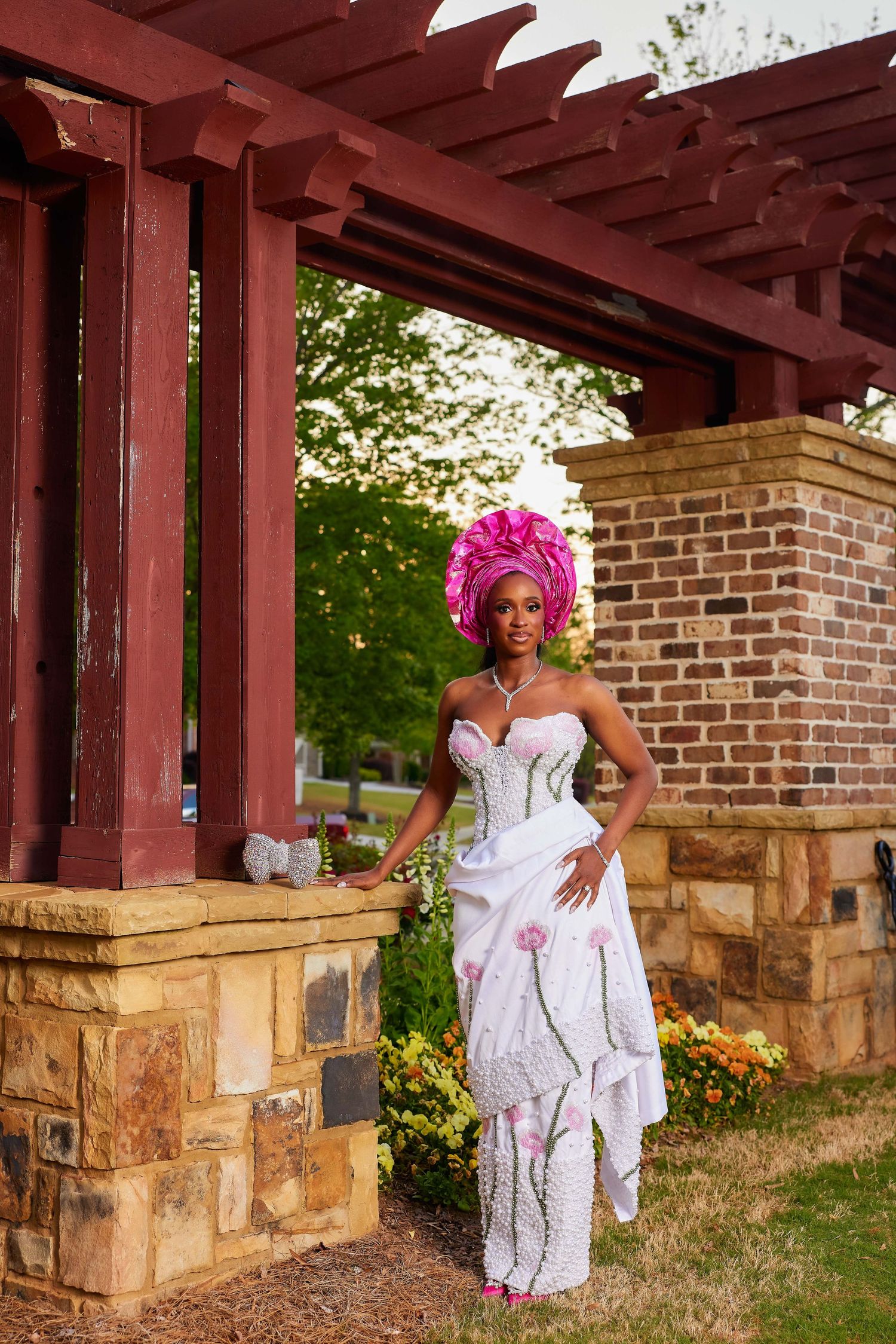 A woman in a white strapless gown and pink headpiece poses under a red pergola in a lush garden setting.