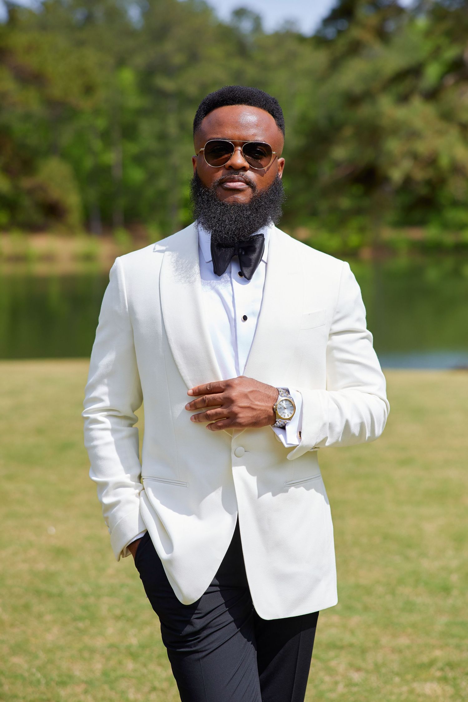 Stylish Black man in white tuxedo jacket, bow tie, sunglasses, and watch poses outdoors near a lake.