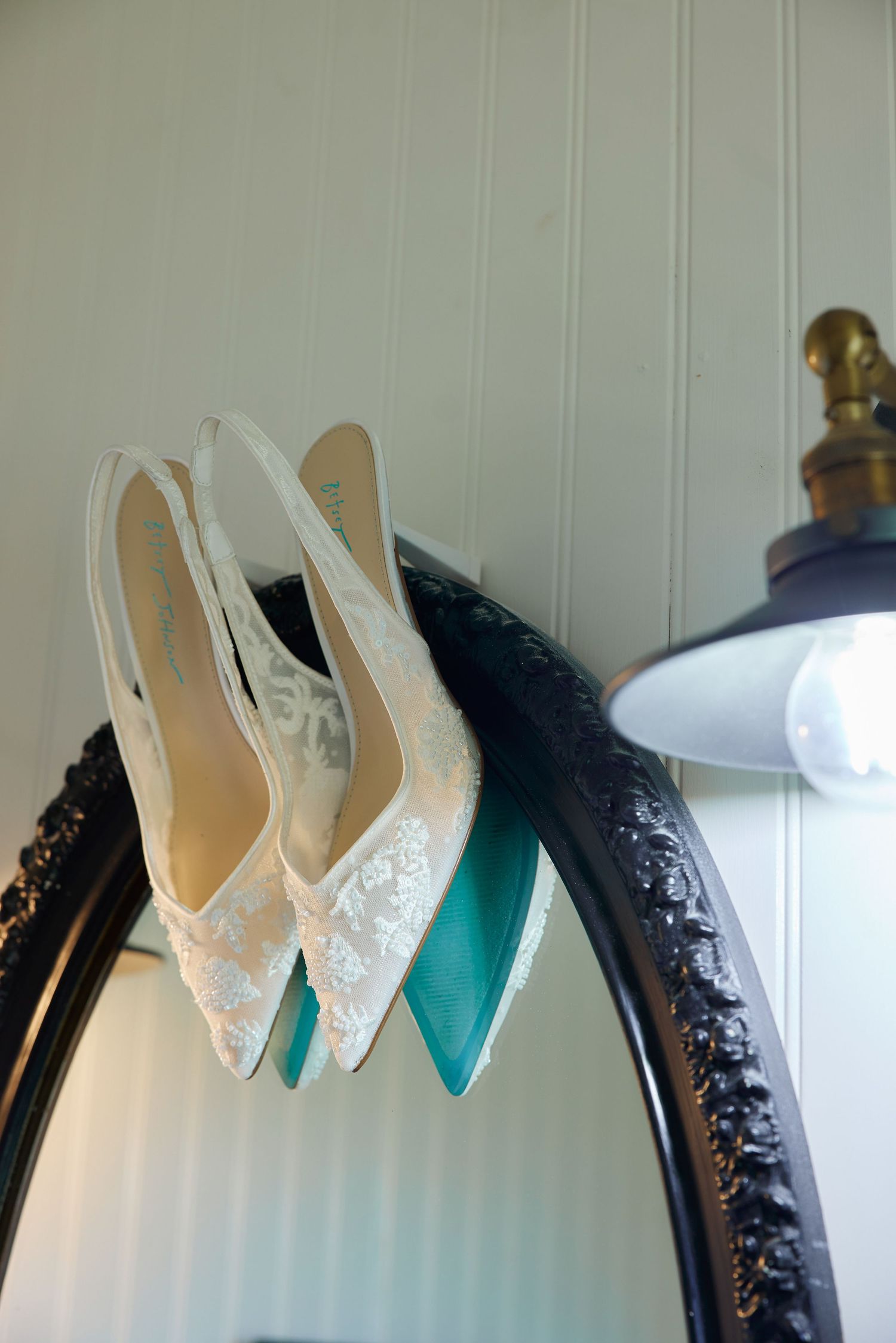 White lace and teal satin bridal heels displayed on an ornate black mirror frame with a lamp in the background.
