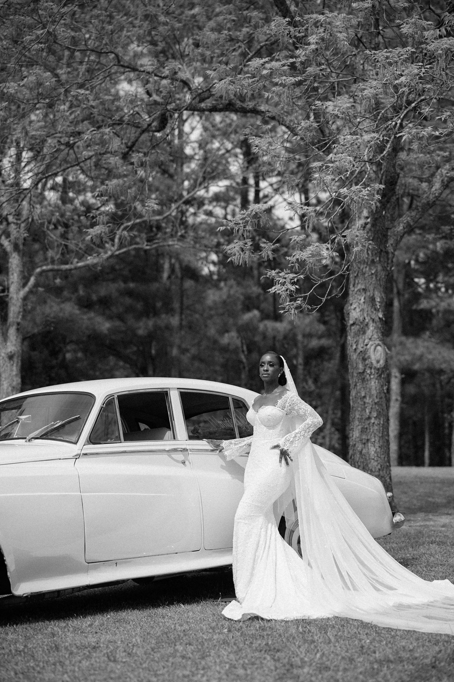 Black and white photo of a bride in a flowing gown posing beside a vintage car under trees.