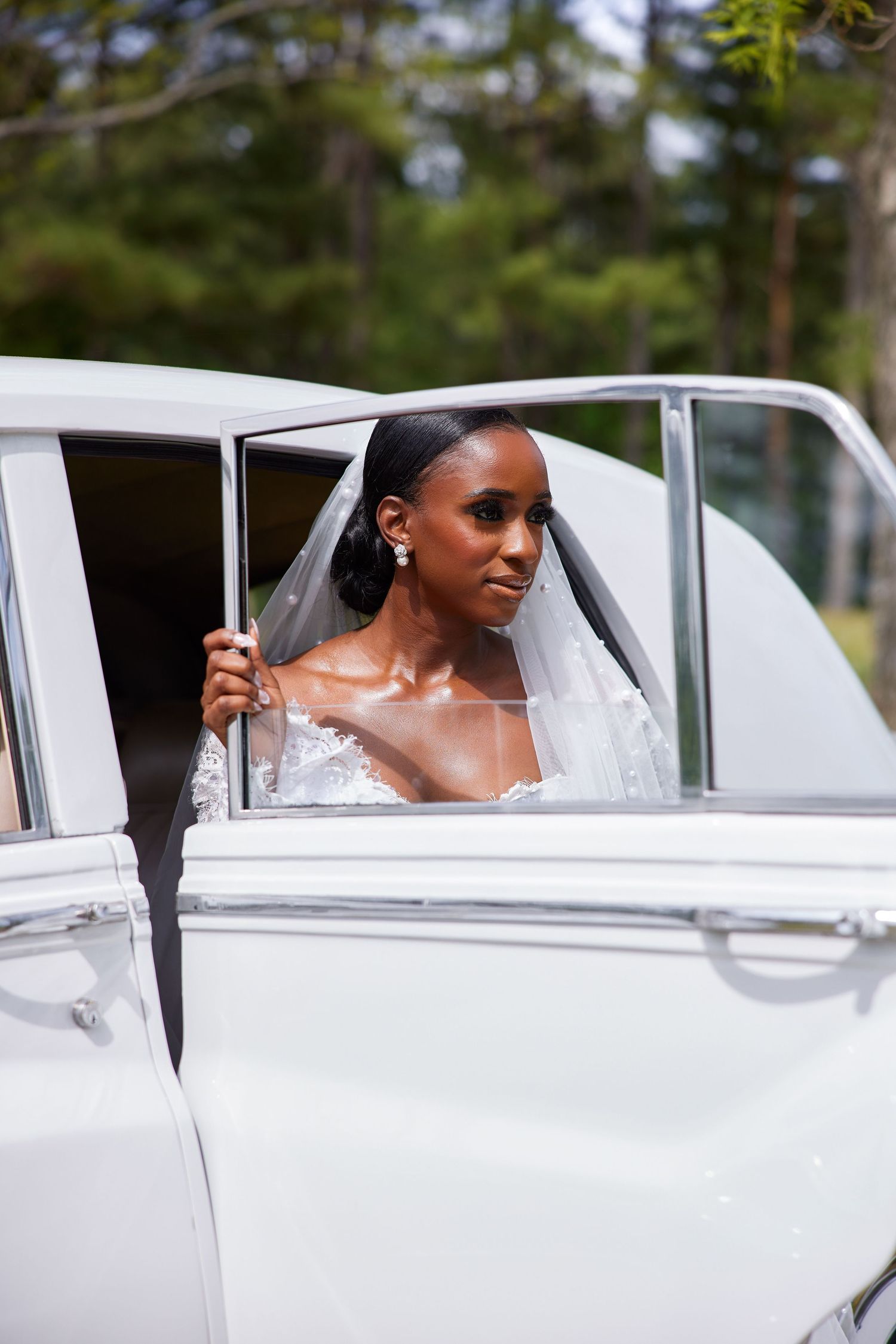 Elegant Black bride in lace wedding gown and veil stepping out of a vintage white car outdoors.