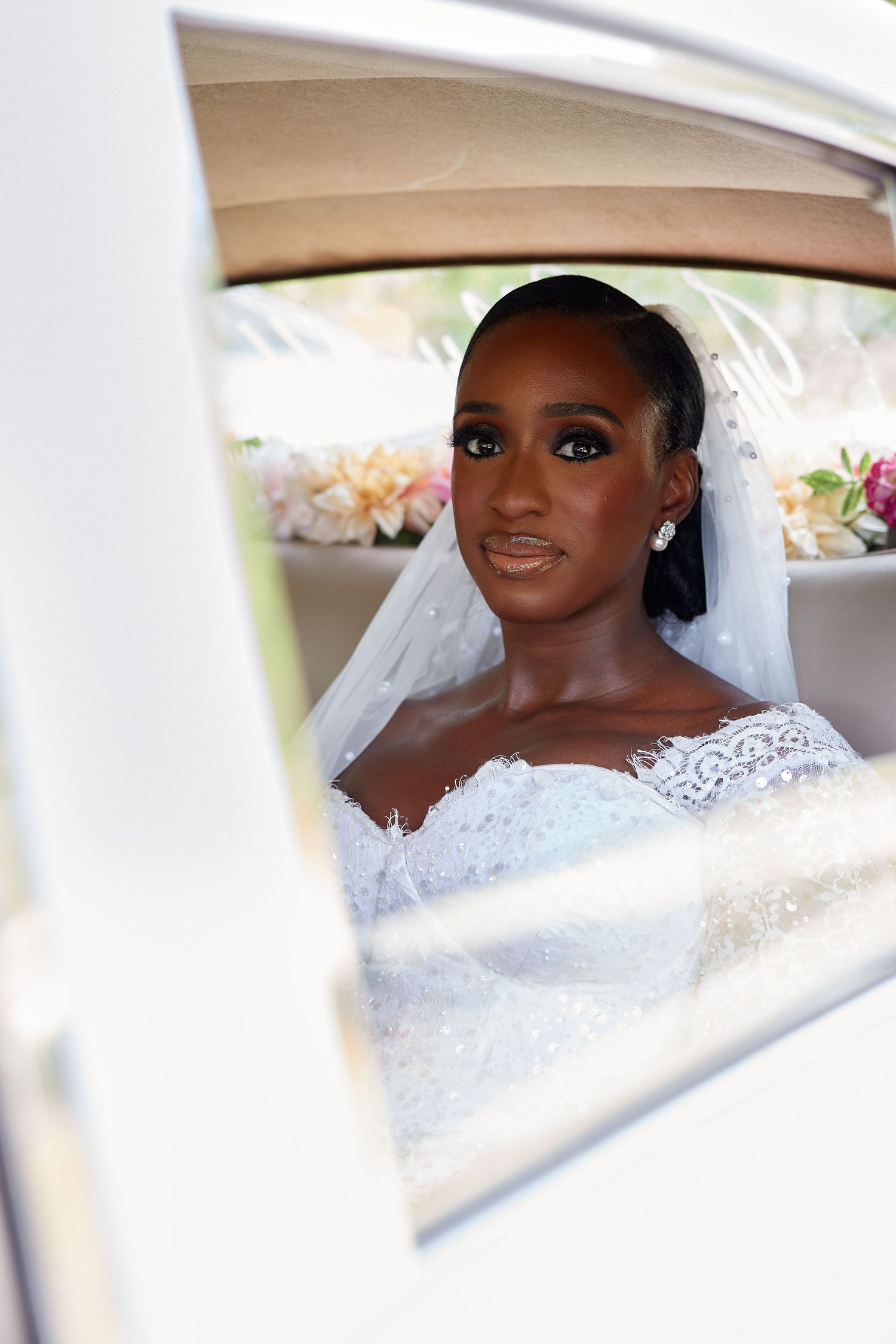 Elegant bride in lace off-shoulder gown and veil seated in a car, looking gracefully through the window.