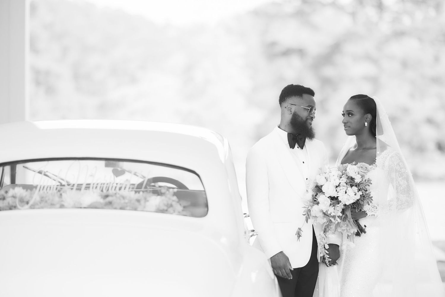 Black and white photo of a bride and groom standing together near a vintage car on their wedding day.